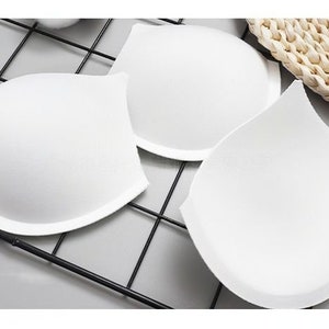Op de afbeelding: Drie witte bh-cups van schuim, gerangschikt op een zwart draadgaas. De cups zijn halfrond en hebben een glad, licht getextureerd oppervlak.