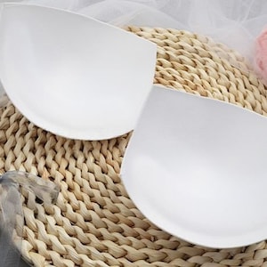 Könnte beinhalten: Zwei weiße Schaumstoff-BH-Einlagen auf einer geflochtenen Strohoberfläche.