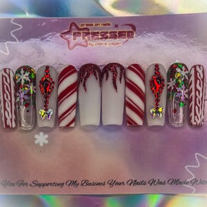 May include: Set of ten press-on nails with a Christmas theme. The nails feature a variety of designs, including candy canes, snowflakes, and a red and white patterned design. The text "PRESSED" is visible in the background.