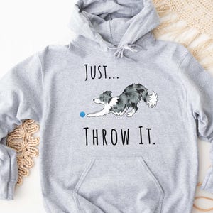 May include: A light gray hoodie with a front pocket and drawstring hood. The hoodie features a graphic of a dog reaching for a blue ball, with the text "Just... Throw It."