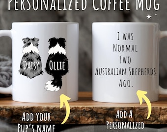 May include: Two white coffee mugs with black and white illustrations of Australian Shepherd dogs. One mug has the text "Daisy" and "Ollie" on it. The other mug has the text "I was normal two Australian Shepherds ago."