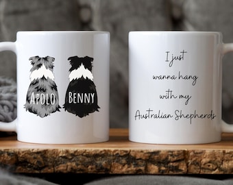 May include: Two white ceramic mugs with black handles. One mug features a black and white Australian Shepherd silhouette with the name "Benny" and the other mug features a gray and white Australian Shepherd silhouette with the name "Apollo". The second mug has the text "I just wanna hang with my Australian Shepherds".