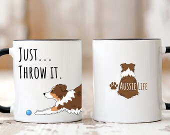 May include: Two white ceramic mugs with black rims. The mug on the left has a brown and white Australian Shepherd dog laying down with a blue ball and the text "JUST...THROW IT." The mug on the right has a brown and white Australian Shepherd dog sitting with its back to the viewer and the text "Aussie Life".
