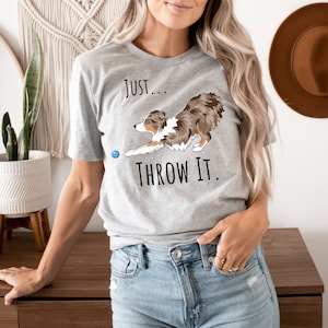 May include: A light grey t-shirt with the text "Just... Throw It." features a cartoon dog reaching for a blue ball. The dog is brown, white, and tan. The shirt is a crew neck.