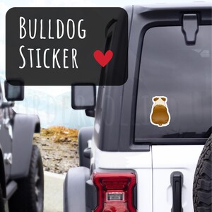 May include: A brown and white bulldog sticker is affixed to the rear window of a white SUV. The sticker is a cartoon illustration of a bulldog's rear end. Above the sticker, a black rectangle displays the words "BULLDOG STICKER" in white, with a red heart.