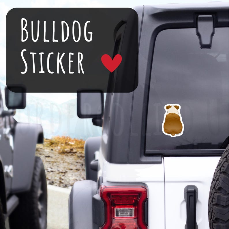 May include: A brown and white bulldog sticker is affixed to the rear window of a white SUV. The sticker is a cartoon illustration of a bulldog's rear end. Above the sticker, a black rectangle displays the words "BULLDOG STICKER" in white, with a red heart.