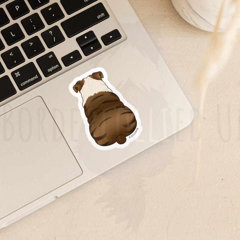 May include: A sticker featuring a brown and white bulldog from the rear. The sticker is on a laptop, with the keyboard visible. The bulldog sticker is a cartoon illustration.