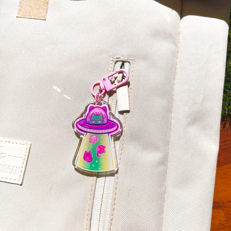 May include: A clear acrylic keychain featuring a cat in a purple UFO with pink and green accents. The keychain is attached to a pink clasp and zipper on a beige backpack. The design includes other cat-shaped aliens.