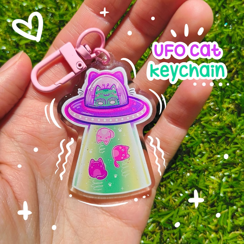 May include: A clear acrylic keychain featuring a purple and pink UFO with cat illustrations. The keychain has a pink clasp and the text "UFO cat keychain" in a playful font. The UFO has a green and pink gradient.