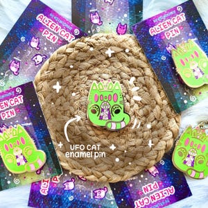 May include: A lime green enamel pin shaped like a cat with a UFO graphic on its chest, featuring pink accents and a striped tail. The pin is displayed on a backing card labeled ALIEN CAT PIN with a space-themed background and white text.