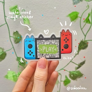 May include: A waterproof vinyl sticker featuring a cartoon illustration of two Nintendo Switch consoles with cat ears. The left console is blue and the right console is red. The screen of the left console shows a pattern of cats with the word "PLAY" in green. The sticker has the text "click" and "waterproof vinyl sticker" on it.