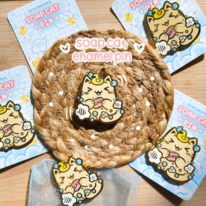 May include: Gold enamel pin of a cartoon cat taking a bath. The cat is holding a bar of soap and is surrounded by bubbles. The pin is on a brown woven background. The text "soap cat enamel pin" is written in white on a pink background.