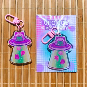 May include: A pink keychain featuring a UFO design with a cat inside the spaceship. The UFO has a purple top and a green and pink interior. The keychain also has pink cat-shaped aliens and the text "UFO Cat Keychain".
