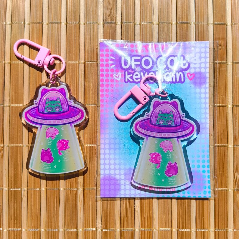 May include: A pink keychain featuring a UFO design with a cat inside the spaceship. The UFO has a purple top and a green and pink interior. The keychain also has pink cat-shaped aliens and the text "UFO Cat Keychain".