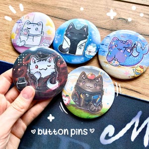 May include: Five circular button pins featuring colourful cartoon cat illustrations. One pin shows a cat in a hoodie, another a cat in a lab coat, a cat mermaid, a cat in a leather jacket with a bird, and a cat with an apple on its head.