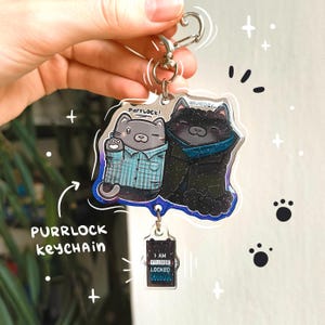 May include: A clear acrylic keychain featuring two cartoon cats, one gray and one black, with the text "PURRLOCK keychain". The gray cat wears a blue plaid shirt, and the black cat wears a blue scarf. A smaller charm reads "I AM PURR LOCKED".
