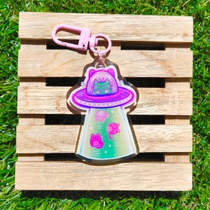 May include: A clear acrylic keychain featuring a pink UFO with a cat inside. The UFO has a pink and purple design with three pink cats floating below. The keychain has a pink clasp.