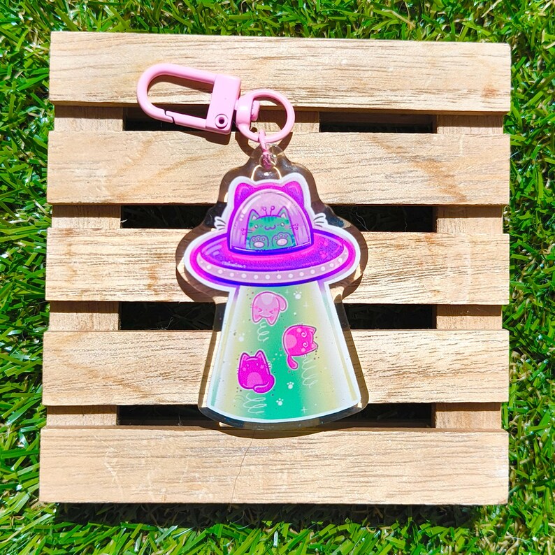 May include: A clear acrylic keychain featuring a pink UFO with a cat inside. The UFO has a pink and purple design with three pink cats floating below. The keychain has a pink clasp.