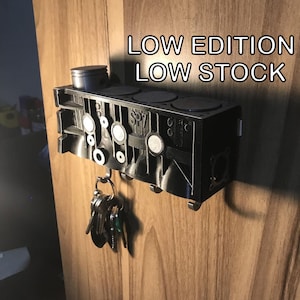 Könnte beinhalten: Schwarzer Schlüsselhalter aus Metall in Form eines Motorblocks mit dem Text "LOW EDITION LOW STOCK" an einer Holzwand.