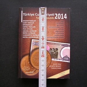 Op de afbeelding: Een bruin boek met de titel "Türkiye Cumhuriyeti 2014" met een liniaal die 40,6 cm meet. De omslag van het boek toont een gouden munt en een Turks bankbiljet met het nummer 10.