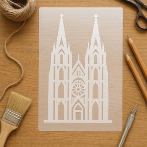Könnte beinhalten: Eine weiße Schablone mit einem detaillierten Architekturdesign einer Kathedrale mit zwei Türmen, einem Rosettenfenster und gewölbten Türen. Die Schablone liegt auf einer Holzoberfläche, mit Kunstbedarf wie Bleistiften, Pinsel und Schere.