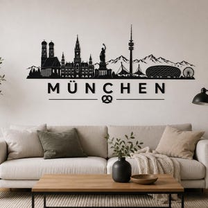 May include: Black metal wall art depicting the Munich skyline, including iconic landmarks like the Frauenkirche, the Olympic Tower, and the Allianz Arena. The word "München" is displayed below the skyline, with a pretzel symbol. The art is mounted on a white wall above a sofa.