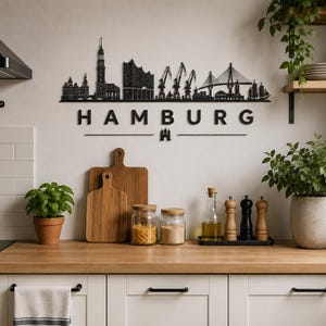 May include: Black metal wall art depicting the Hamburg skyline, with the city name below. The kitchen setting includes wooden cutting boards, glass jars, and a black stovetop. Green plants and kitchen utensils add color.