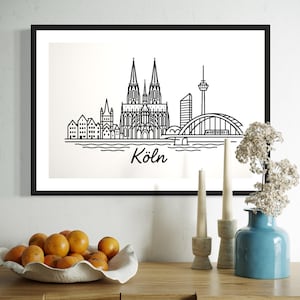 May include: Black framed print featuring a minimalist line drawing of the Cologne skyline, including the cathedral, bridge, and TV tower. The word "Köln" is written below the image. The print is displayed on a white wall.