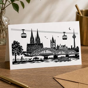 May include: A white greeting card featuring a black silhouette illustration of the Cologne skyline. The design includes the cathedral, the Hohenzollern Bridge, a cable car, a boat on the river, a cyclist, and people sitting on a bench.
