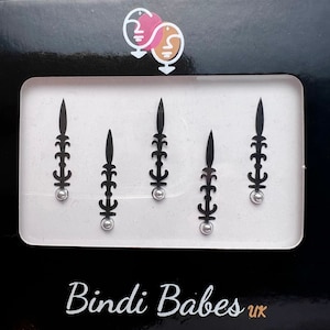 Puede incluir: Un paquete de cinco pegatinas bindi decorativas negras y plateadas. Las pegatinas tienen forma de flecha estilizada con una pequeña gema plateada en la parte inferior. El paquete está etiquetado como "Bindi Babes UK".