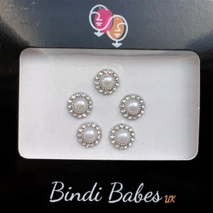 May include: A package of five decorative bindis. Each bindi features a central faux pearl surrounded by a ring of small, clear rhinestones. The package is black with the words "Bindi Babes uk" in white.