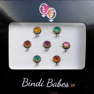 Può includere: Un set di sette bindis colorate con gioielli e dettagli dorati. Le bindis sono disposte in una griglia su uno sfondo bianco. Le bindis sono confezionate in una scatola nera con il testo "Bindi Babes uk" stampato sul fondo.