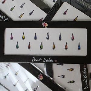 May include: A rectangular black display card labeled Bindi Babes UK featuring a row of twelve adhesive bindi stickers. Each bindi is shaped like a pointed teardrop with a serrated edge, presented in various colors including yellow, green, red, blue, and purple.