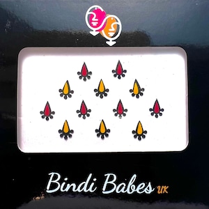 May include: A package of decorative bindis in a white tray, set against a black background. The bindis are teardrop-shaped with red and yellow centers, surrounded by black dots. The text "Bindi Babes uk" is printed at the bottom.