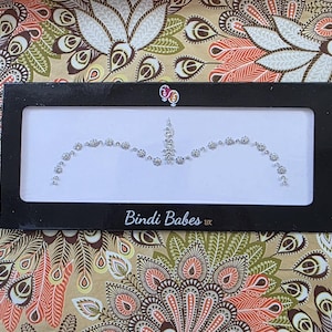 May include: A black rectangular box with a white interior displays a silver forehead jewel. The jewel is in an arch shape, composed of small, round, sparkling gems. The text "Bindi Babes" is printed on the box.
