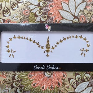 Könnte beinhalten: Ein dekoratives, goldfarbenes Stirn-Bindi-Design mit einem zentralen vertikalen Element und geschwungenen Linien aus kleineren Verzierungen. Das Bindi wird in einer schwarzen Box mit dem Text "Bindi Babes uk" präsentiert.