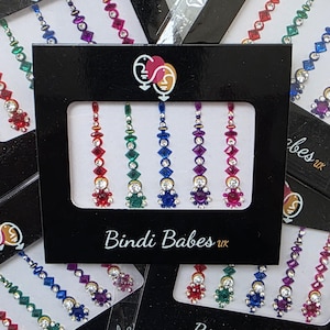 May include: A collection of colorful bindi forehead jewels in various colors, including red, green, blue, and purple. Each jewel features a central design with surrounding rhinestones. The packaging displays the text "Bindi Babes UK".