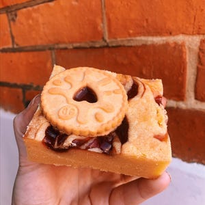 May include: A square-shaped blondie bar topped with a round, light-colored biscuit featuring a central heart-shaped cutout. The blondie has a golden-yellow base with visible swirls of dark red fruit jam or preserves embedded throughout the baked dough.