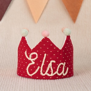 K&ouml;nnte beinhalten: Rote Filzkrone mit wei&szlig;en Punkten und wei&szlig;er Stickerei, die "Elsa" sagt. Die Krone hat drei wei&szlig;e Bommeln oben, einen rosa, einen gr&uuml;nen und einen blauen.