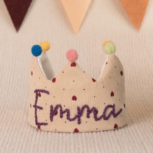 K&ouml;nnte beinhalten: Eine beige Krone mit roten Punkten und einem in Lila gestickten Namen "Emma". Die Krone ist mit bunten Bommeln verziert.