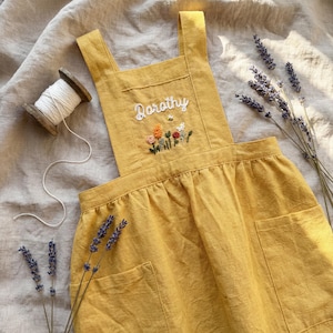 以下が含まれることがあります： マスタードイエローのリネンジャンパースカート。刺繍された花のディテールと「Dorothy」という名前が特徴です。ドレスには2つのフロントポケット、スクエアネックライン、調節可能なストラップが付いています。白い糸の糸巻きとラベンダーの小枝も画像にあります。