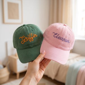 May include: A dark green baseball cap with the name Griffin embroidered in orange thread and a light pink baseball cap with the name Eleanor embroidered in purple thread, both featuring a structured crown and a curved brim.