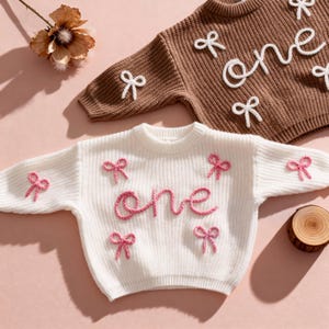 Può includere: Sono mostrati due maglioni per bambini lavorati a maglia, uno marrone e uno bianco. La parola "one" è ricamata su ogni maglione. Il maglione bianco ha fiocchi rosa e la parola "one" in rosa. Il maglione marrone ha fiocchi bianchi e la parola "one" in bianco.