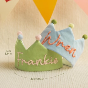 K&ouml;nnte beinhalten: Zwei Stoffkronen mit gestickten Namen. Die gr&uuml;ne Krone sagt "Frankie" in rosafarbenem Garn und die blaue Krone sagt "Wren" in orangefarbenem Garn. Die Kronen sind 8 cm hoch und 30 cm im Umfang.