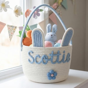 以下が含まれることがあります： 青い持ち手と「Scottie」という名前が青い文字で書かれた、白いロープのイースターバスケット。バスケットには、ぬいぐるみウサギ、ニンジンのおもちゃ、装飾用の卵が入っています。前面には青い花が飾られています。
