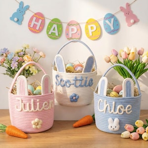 Könnte beinhalten: Drei Ostkörbe in Rosa, Weiß und Blau, jeweils mit Hasenohren und personalisierten Namen. Die Körbe sind mit Heu und bunten Eiern gefüllt, mit einer Karotte und einem "Happy Easter" Banner im Hintergrund.