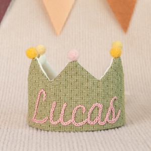 K&ouml;nnte beinhalten: Gr&uuml;ne Filzkrone mit rosa Filzstichen, die den Namen "Lucas" buchstabieren, und gelben Filzpompons oben.