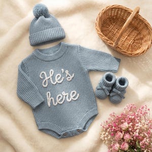 Op de afbeelding: Een lichtblauwe gebreide romper met de tekst "He's here" in witte letters, een bijpassende muts met pompon en babyslofjes. Een rieten mand en een boeket roze bloemen zijn ook in de afbeelding te zien. De babykleding ligt op een crèmekleurige deken.