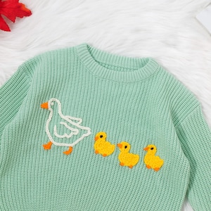 May include: A light green knitted sweater with a white embroidered goose and four yellow embroidered ducklings.