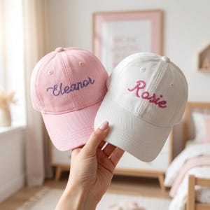 May include: A light pink baseball cap with the name Eleanor embroidered in purple thread and a white baseball cap with the name Rosie embroidered in pink thread. Both caps feature a structured crown, a curved brim, and a button on the top.